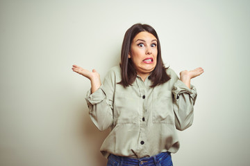 Young beautiful brunette woman wearing green shirt over isolated background clueless and confused expression with arms and hands raised. Doubt concept.