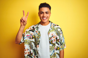 Young brazilian man on vacation wearing summer floral shirt over isolated yellow background showing and pointing up with fingers number two while smiling confident and happy.