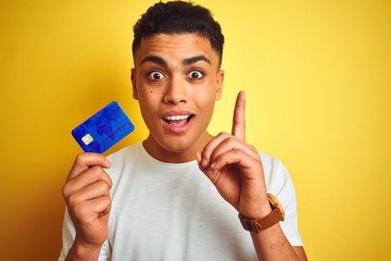 Young brazilian customer man holding credit card standing over isolated yellow background surprised...