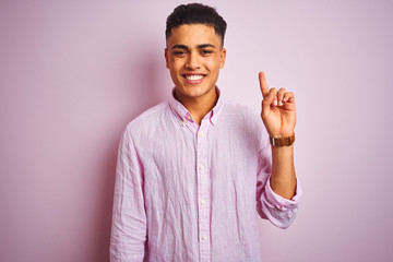 Young brazilian man wearing shirt standing over isolated pink background showing and pointing up with finger number one while smiling confident and happy.