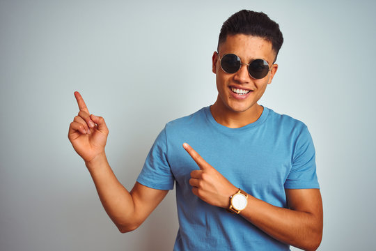 Young brazilian man wearing blue t-shirt and sunglasses over isolated white background smiling and looking at the camera pointing with two hands and fingers to the side.