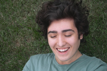 The white-toothed curly guy lies on the green grass and laughs with his eyes closed