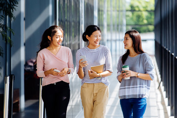Young Asian business women talking while walking in office building in casual wear. Three girls strolling and showing colleague her phone outdoors