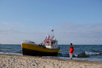 Wooden fishing boat on the beach of Baltic Sea in Sopot/Poland. Silhouette of little boy in red jacket standing near boat with raised hands and looking with delight to waves.