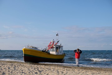 Naklejka na ściany i meble Wooden fishing boat on the beach of Baltic Sea in Sopot/Poland. Silhouette of little boy in red jacket standing near boat with raised hands and looking with delight to waves.