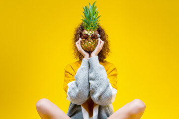 Curly slim girl is holding pineapple dressed in glasses in front of her face. Sunny young female is sitting on yellow background. Bright active lifestyle. Good mood and fun pastime concept.