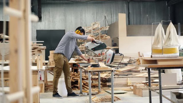Male worker in headphones and goggles is using electric saw in wood workshop sawing timber working alone focused on activity. Business and woodwork concept.