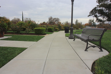 Community park. Sidewalk leading between green grass, bushes, lampstand & bench.