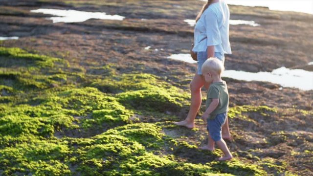 Slow motion of two years old boy walking with his mother on green algae at low tide in the evening. Balangan beach, Bali, Indonesia