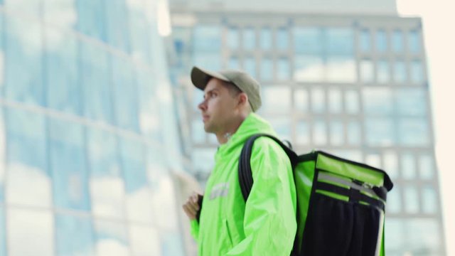 Zoom out tracking left shot of young food courier in green uniform and cap walking down city street with insulated backpack. Man passing by modern glass building delivering order, side view