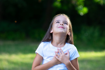 Little girl looking up to at sky with hands on chest, summer nature outdoor. Happy smiling kid...