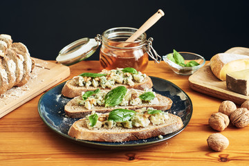 Close up on a plate of toasts with roquefort cheese, walnuts, honey and basil. Some cheese, nuts and honey in the background