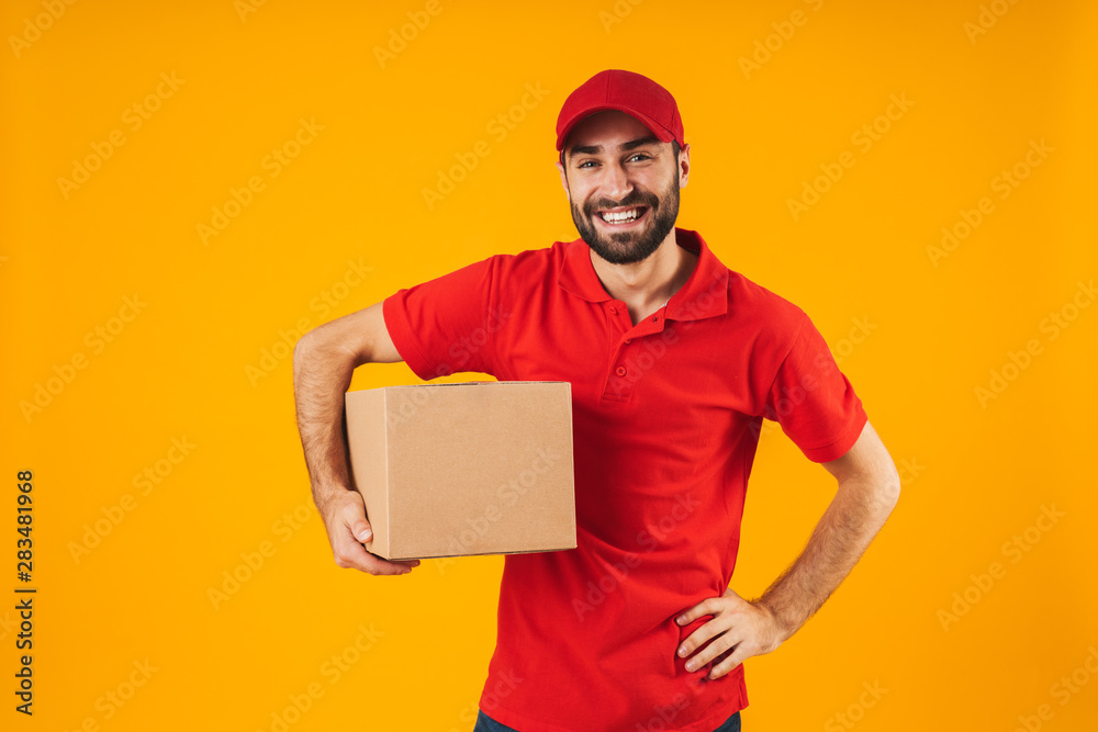 Sticker Portrait of cheerful delivery man in red uniform smiling and holding packaging box