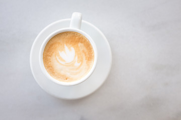 Cup of coffee with milk on marble table with wooden chairs in unfocused background, top view