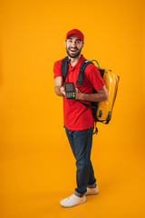 Photo of cheerful delivery man in red uniform holding payment terminal while carrying backpack with takeaway food
