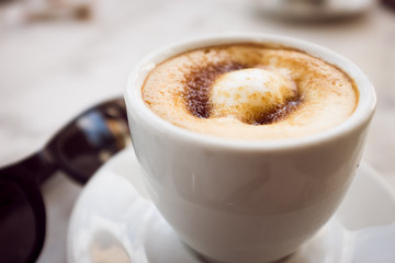 Cup of coffee with milk and brown sugar on marble table with sunglasses in the background