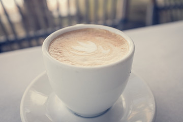 Cup of coffee with milk on marble table with wooden chairs in unfocused background, vintage style