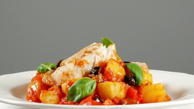 Traditionally prepared cod in Spanish. Served in tomato sauce with boiled potatoes, olivki and basil. Front view.  Food presentation. Turning the platter.