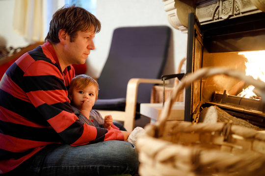Middle age father sitting by fireplace with cute little baby girl at home. Happy family, dad with daughter make fire for Christmas time. Safe handling for kids - Powered by Adobe