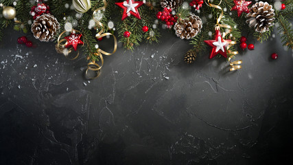 Festive Christmas greeting layout template with fir branches decorated with natural fir cones, gold serpentine, red berries and stars. Dark black textured background, copy space, top view.