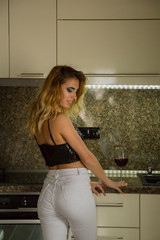 Young blonde woman in sparkling top and white jeans posing in kitchen