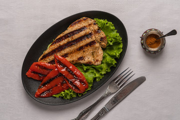 Grilled turkey breast steak with baked peppers and lettuce leaves in a oval black plate with knife and fork on a light linen background. Next to the pepper shaker with red pepper. Top view, copy space