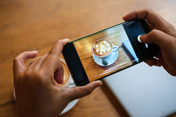 Hand taking a picture of hot latte art coffee cup with art foam on wooden table with her smart phone sitting at a coffee shop snap and share on social media by smartphone. Web Blogger photo shoot 