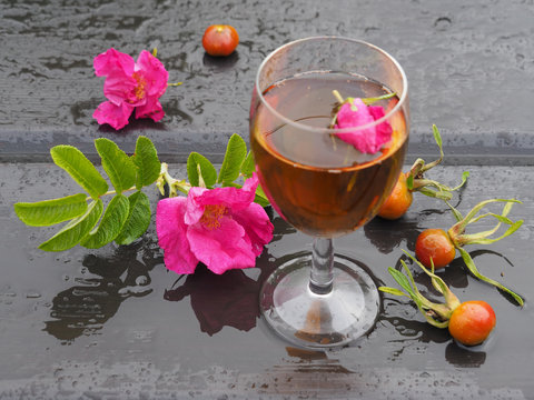 Wine in a transparent glass of a healthy drink and rosebud. Goblet with a beautiful tincture, rose flower and berries on a wet table in the rain. Original picture for the wishes of good health.