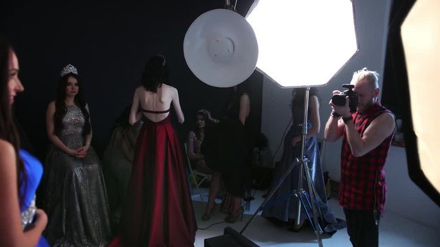 man photographer, photographing a girl model, brunette, in a blue long dress with a deep neckline, beautiful hair and bright makeup, with a crown, in the Studio, on a black background, a lot of girls