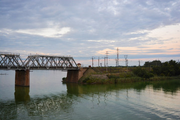 The gray Samara railway bridge over the surface of the Samara river with a beautiful landscape, green trees, bushes and greenery. Pictures of the river landscape with an unusual sky and bright clouds.