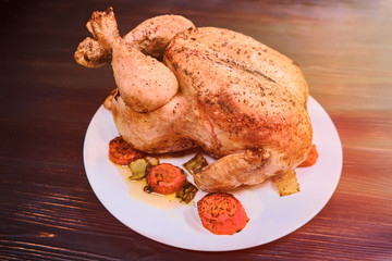 Whole grilled chicken with golden crust on a plate. Fried chicken on redwood background, mahogany. Festive dinner with baked bird.