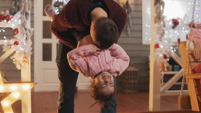 Happy father having fun with her kid daughter at Christmas porch. Dad tickling her little daughter in slow motion. Father and daughter in pink pullover at Christmas day. Close-up shot