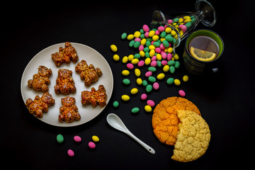 Homemade bear shape candies with honey, sesame and sunflower seeds,glass vase with colorful candies and cup of tea with lemon slice on a black background. Sweet breakfast or snack.