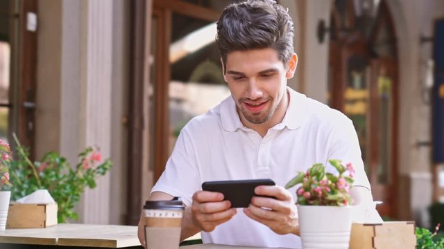 Young happy handsome man sitting in cafe outdoors at the street play games by mobile phone drinking coffee
