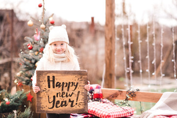 Smiling baby girl 4-5 year old holding table with Happy New Year sign over lights closeup. Celebration.