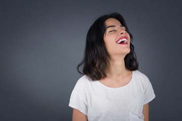 Joyful European woman having fun and laughs at good joke or sings wears casual clothes, standing against gray wall. Happy woman with long hair poses inddoors.