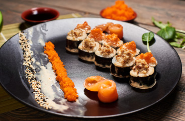 Appetizing hot sushi rolls in creamy sauce serving with salmon, caviar and sesame seeds in big black plate. Sauces and leaves on blurred background. Front detailed view. Exotic Japanese dish recipe.