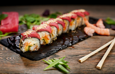 Sushi covered with tuna and sesame seeds on sloppy lubricate stone plate. Focus on details. Front view. Bamboo chopsticks, green onion on table. Tuna piece, shrimps, salad leaves on blurred background