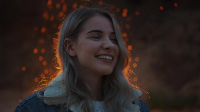 Portrait of a Young Beautiful Blond Woman in a Romantic Evening Atmosphere with a Campfire in the Background. She Expresses a Cute Smile. Fire Reflects on Her Hair and She Wears a Barbell Earring.