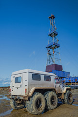 Off-road car with big wheels. White color. Dirty. In the background is a drilling rig. The concept of solving the transport problem in oil production in off-road conditions.