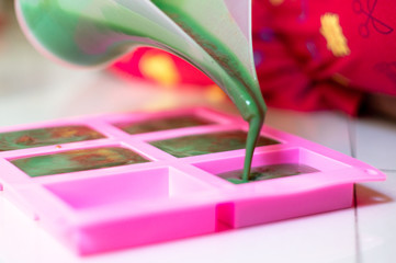 Pouring green colored soap mix with red swirls from a vessle with snout into a pink mold for making...