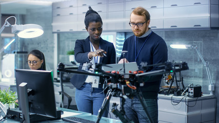 Caucasian Male and Black Female Engineers Working on a Drone Project with Help of Laptop and Taking Notes. He Works in a Bright Modern High-Tech Laboratory.