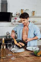 sexy smiling man holding frying pan with fish in kitchen