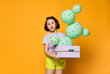 Young brunette girl hold cactus balloon in pastel pink wooden box. Flowers are made of balloons with pink flowers and painted spines. Creative idea minimal concept on yellow background