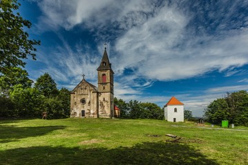 Krasikov, Kokasice / Czech Republic - August 9 2019: View of the old church of Mary Magdalene and a bell tower. Sunny summer day. Green grass, blue sky with white clouds.
