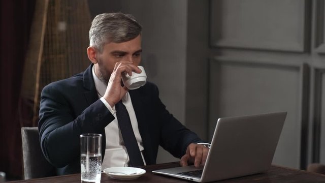 Chest-up shot of 40-something Caucasian male executive in business suit, shirt and tie sitting at table in restaurant meeting room, working on laptop, looking at screen, and drinking coffee and water