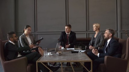 Full dollying shot of five multinational male and female corporate executives in business suits sitting around table in meeting room, passing around document on clipboard, reading and commenting on it