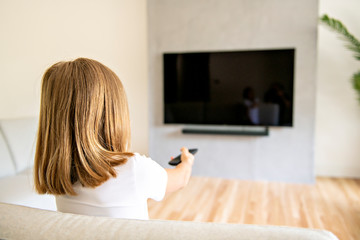 Back view image of cute little blond hair girl sitting on sofa and watching TV.