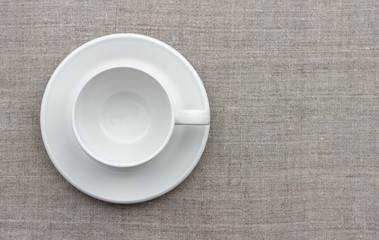 White cup and saucer on natural linen fabric, top view, free space