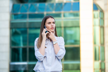 Executive business woman talking on mobile smartphone in the street with office buildings in the background. Young woman with smartphone standing against street blurred building background.